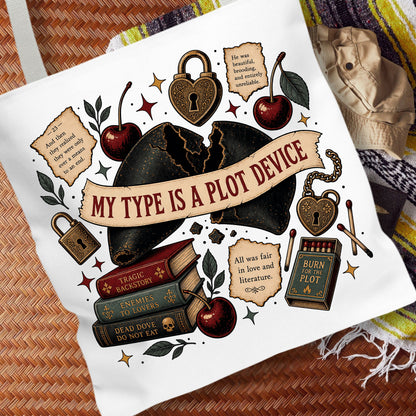 A white canvas tote bag featuring a detailed gothic-themed graphic print with a broken black heart, padlocks, cherries, books, and quote cards. The bag is displayed flat on a woven brown surface next to a beige hat and a striped blanket.