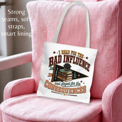 A white canvas tote bag with a vintage-style graphic reading 'I Read for the Bad Influence and Stayed for the Consequences,' styled on a pink armchair with a pink pillow. Text on the left highlights strong seams, soft straps, and smart lining.