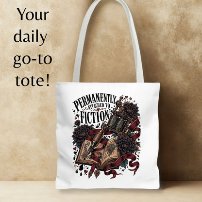 A white canvas tote bag with a gothic-style graphic print featuring an open book, lantern, roses, and the text 'PERMANENTLY ATTACHED TO FICTION'. The bag is shown against a textured beige background with a text overlay on the left reading 'Your daily go-to tote!'.