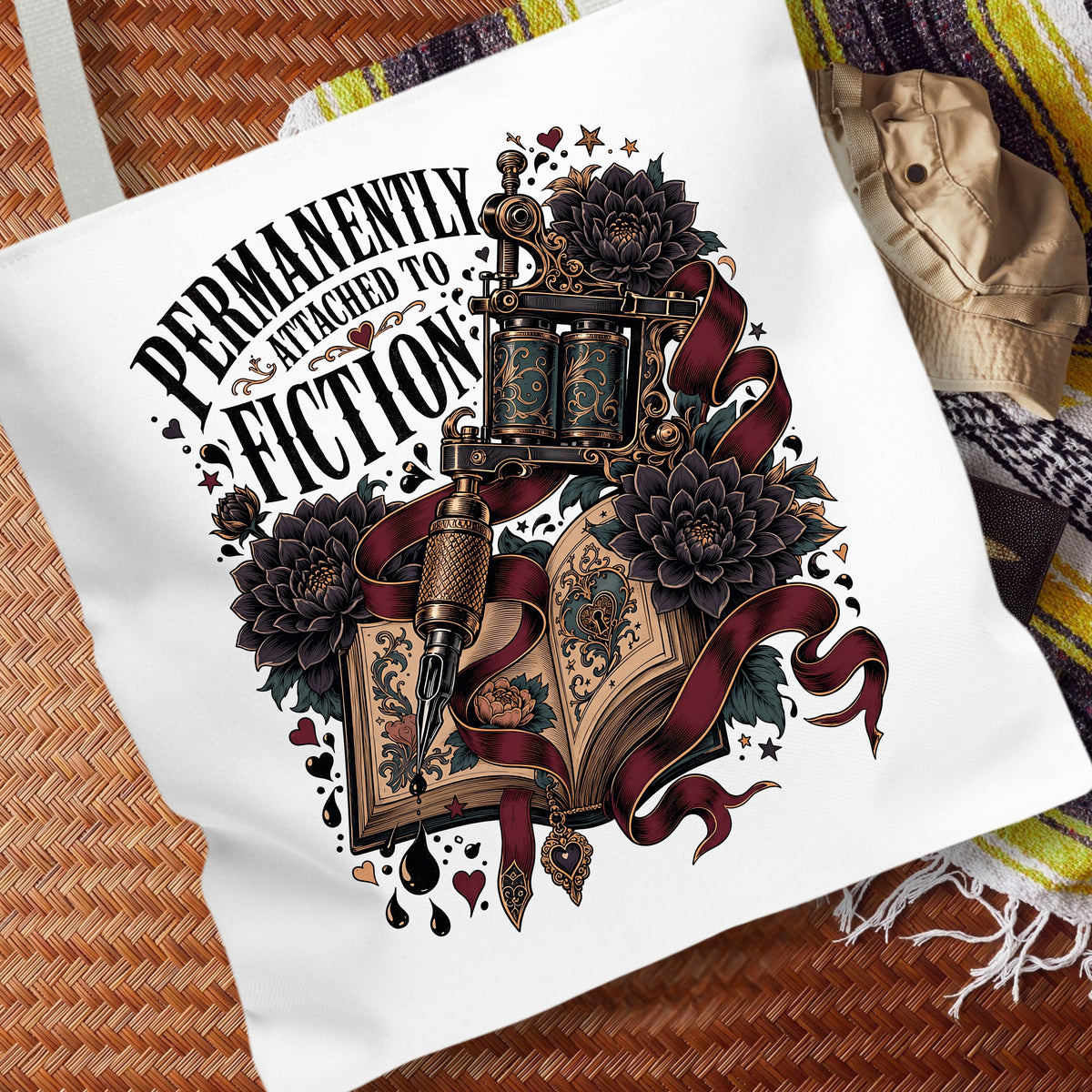 A white canvas tote bag laid flat on a woven brown mat, featuring a large graphic print with the text 'PERMANENTLY ENTHUSIAST TO FICTION' and an illustrated open book, tattoo machine, flowers, and dark ribbons.