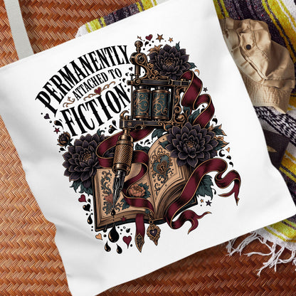 A white canvas tote bag laid flat on a woven brown mat, featuring a large graphic print with the text 'PERMANENTLY ENTHUSIAST TO FICTION' and an illustrated open book, tattoo machine, flowers, and dark ribbons.