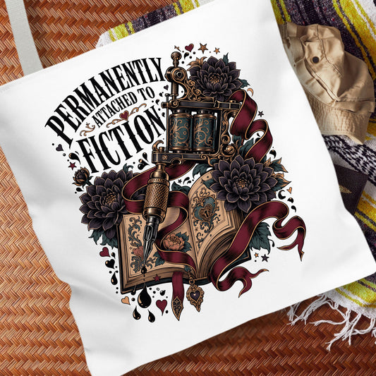 A white canvas tote bag laid flat on a woven brown mat, featuring a large graphic print with the text 'PERMANENTLY ENTHUSIAST TO FICTION' and an illustrated open book, tattoo machine, flowers, and dark ribbons.