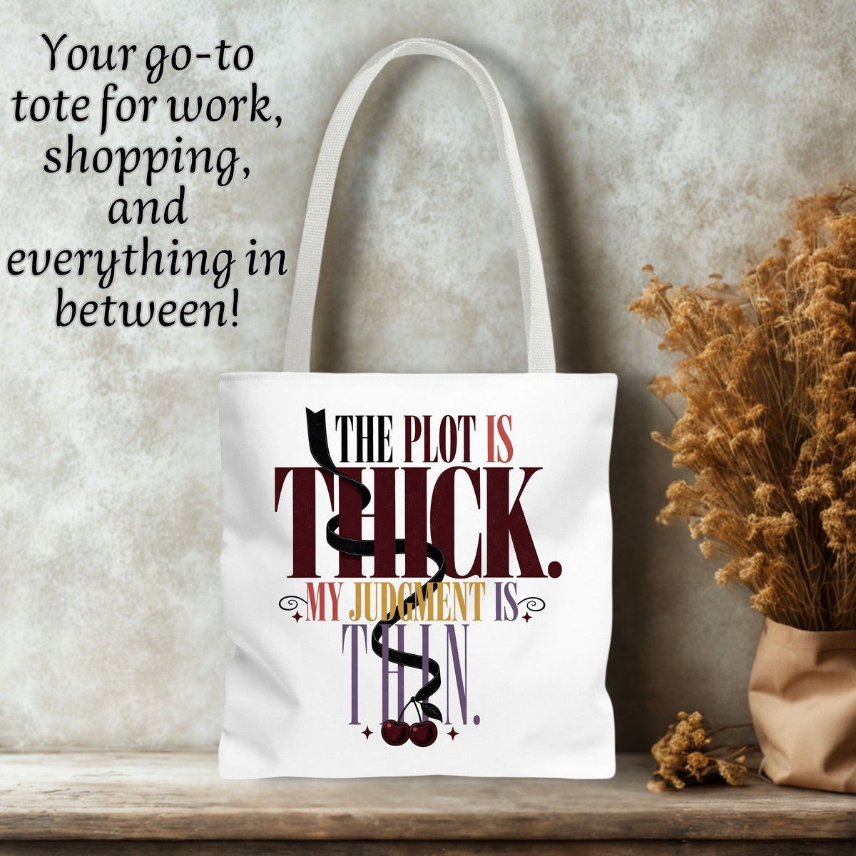 A white canvas tote bag with long handles stands on a wooden surface against a textured gray wall. The bag has a printed quote in dark red, black, and gold text reading 'THE PLOT IS THICK.