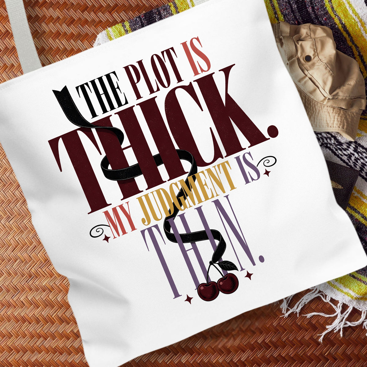 A white canvas tote bag with a humorous quote printed on it, styled on a woven brown mat with a beige hat and a striped blanket nearby.
