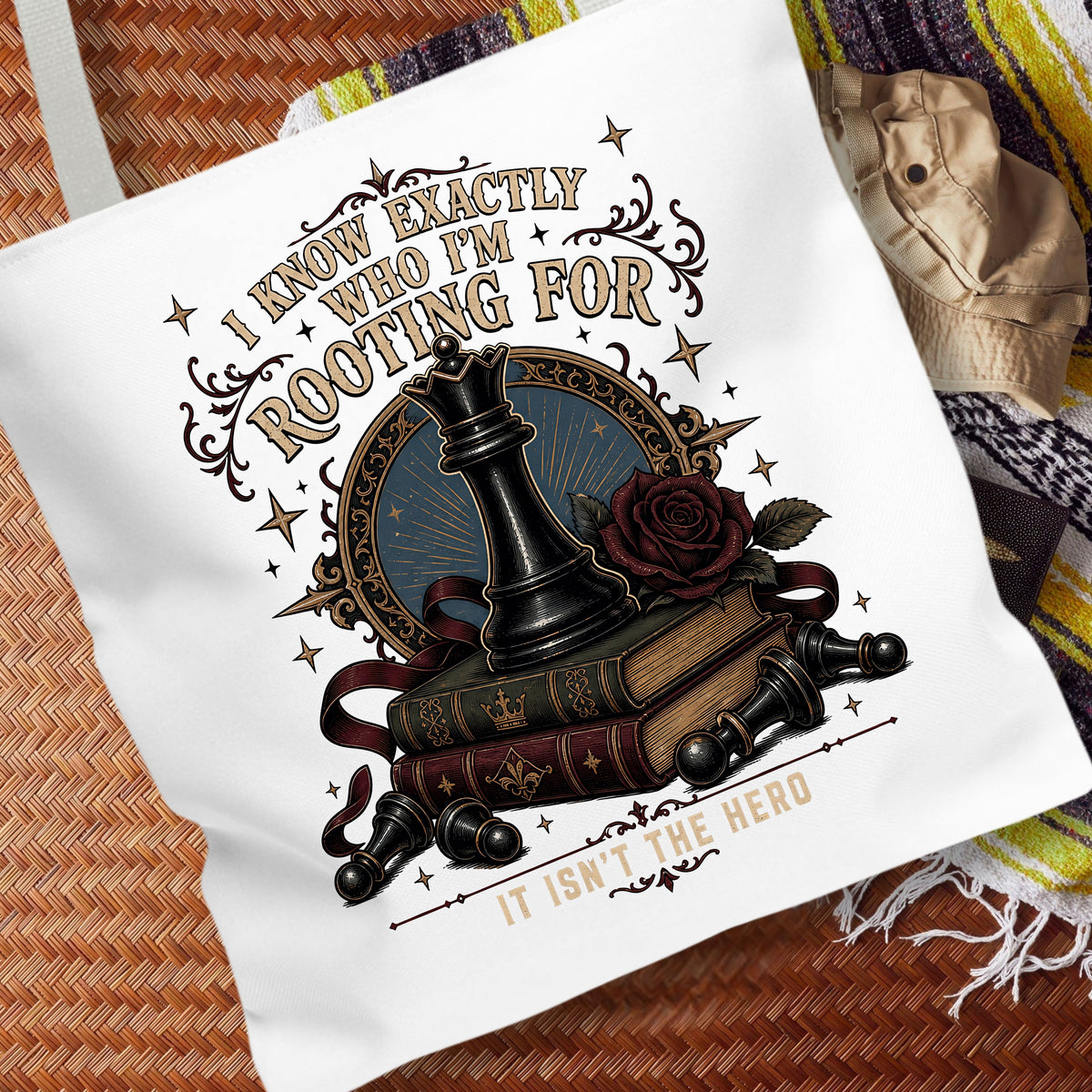 A white canvas tote bag with a detailed graphic print featuring a chess piece, books, a rose, and the text 'I KNOW EXACTLY WHO I'M ROOTING FOR IT ISN'T THE HERO'. The bag is displayed on a woven brown mat with a striped blanket in the background.