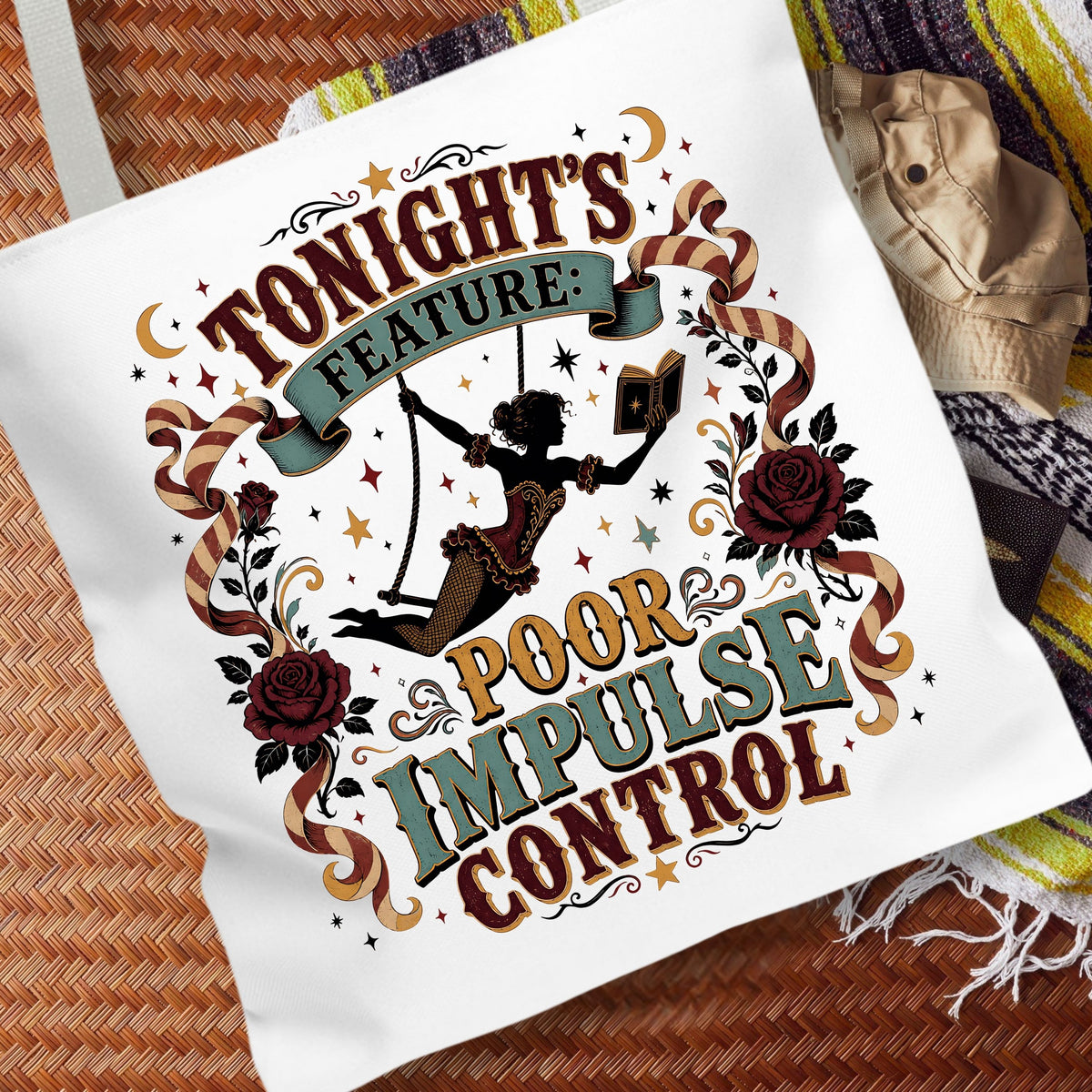 A white canvas tote bag with a vintage-style graphic print reading 'TONIGHT'S FEATURE: POOR IMPULSE CONTROL' in brown, teal, and gold lettering, surrounded by roses, stars, and a swing illustration. The bag is styled on a woven brown mat with a striped blanket and a small hat nearby.