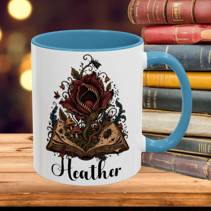 A white ceramic mug with a blue handle and interior, featuring a detailed graphic of a dark red flower blooming from an open book with fantasy creatures, and the name 'Heather' printed below in black script. The mug is styled on a wooden surface next to a stack of vintage books against a warm, blurred background.