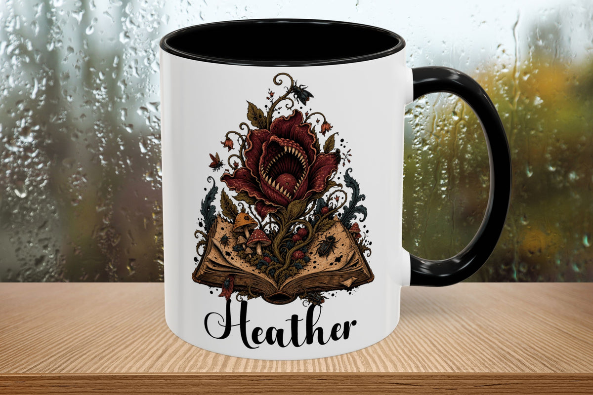 A white ceramic mug with a black handle and black interior, featuring a graphic of a red rose opening an ancient book with the name 'Heather' written below in black script. The mug is placed on a wooden surface with a blurred, rainy window background.