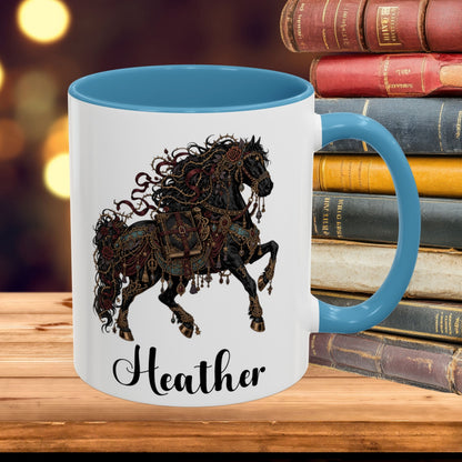 A white ceramic mug with a blue interior and handle, featuring a detailed black and brown steampunk horse illustration and the name 'Heather' in black script. The mug is set on a wooden surface next to a stack of vintage books with a blurred warm background.