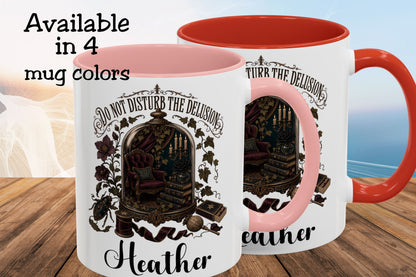 Two white ceramic mugs with pink and red interiors/handles are shown on a wooden surface against a soft beach background. The mugs feature a dark illustrated scene with the text 'DO NOT DISTURB THE DELUSION' and the name 'Heather'.