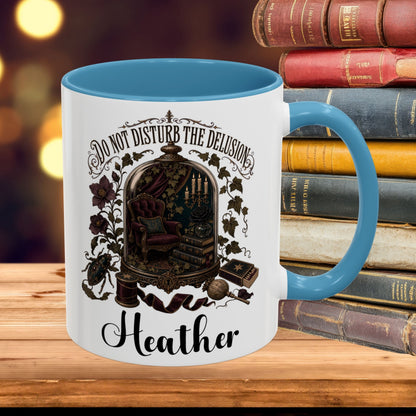 A white ceramic mug with a blue interior and handle, featuring a gothic illustration of a cabaret scene and the text 'DO NOT DISTURB THE DELUSION Heather' on the front. The mug is placed on a wooden surface next to a stack of vintage books with a blurred warm bokeh background.