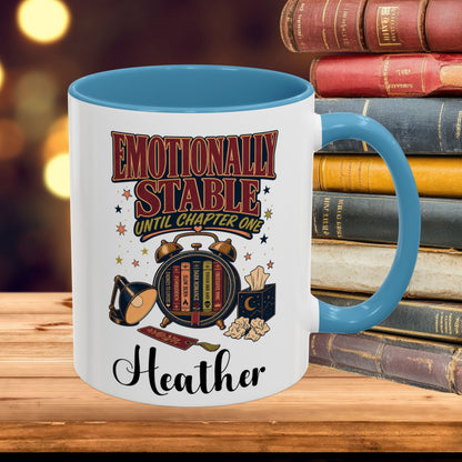 A white ceramic mug with a blue interior and handle, featuring a book-themed graphic that reads 'Emotionally Stable Until Chapter One' and the name 'Heather'. The mug is placed on a wooden surface with a stack of vintage books in the background.