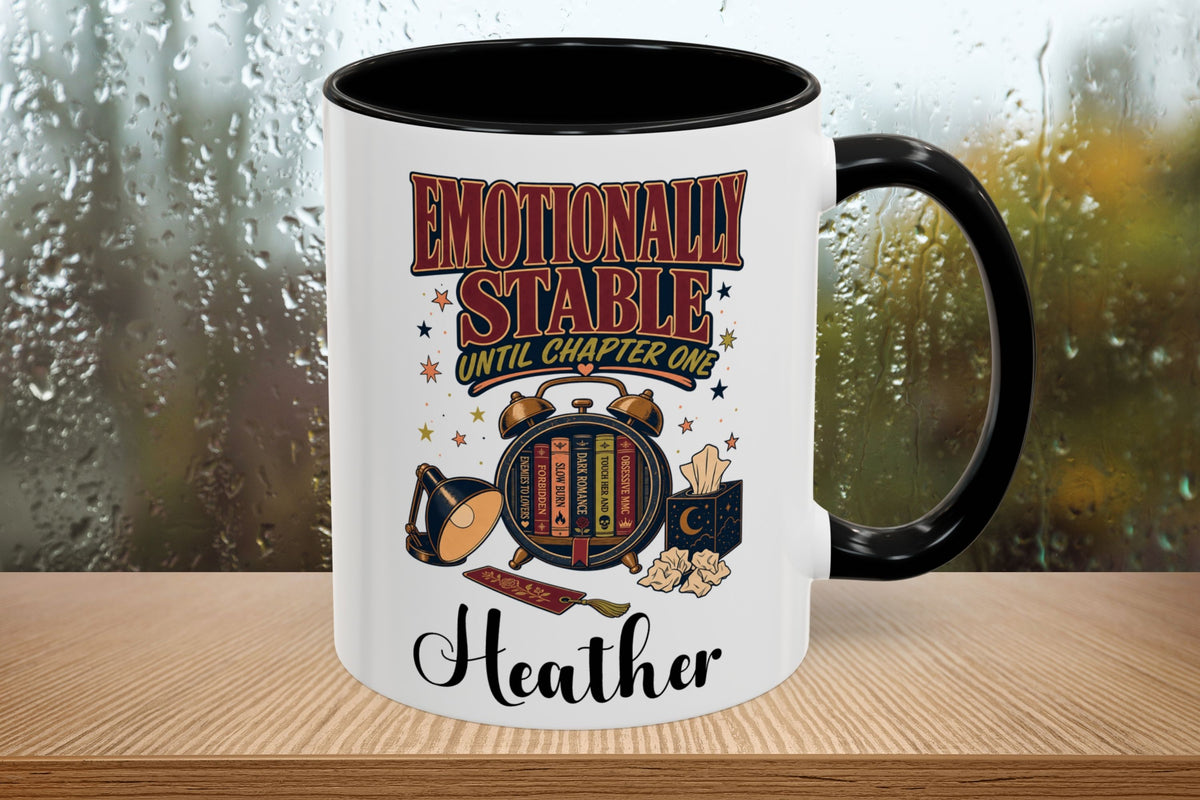 A white ceramic mug with a black interior and handle, featuring a graphic design with the text 'EMOTIONALLY STABLE UNTIL CHAPTER ONE' and the name 'Heather' at the bottom, displayed on a wooden surface with a blurred rainy window background.