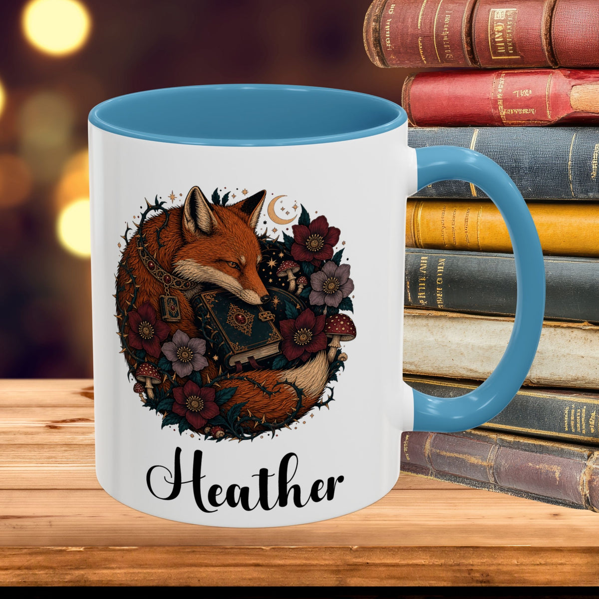 A white ceramic mug with a blue handle and interior, featuring an illustration of a sleeping fox surrounded by flowers and the name 'Heather' printed below. The mug is set on a wooden surface next to a stack of vintage books with a blurred warm background.