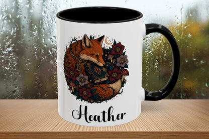 A white ceramic mug with a black handle and black interior, featuring a printed illustration of a fox surrounded by flowers and the name 'Heather' in script below. The mug sits on a wooden surface with a blurred rainy window background.