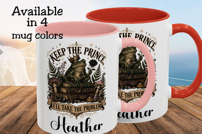 Two white ceramic mugs with red interiors and handles, shown on a wooden surface with a blurred beach background. The front mug has a printed graphic of a frog on a suitcase with the text 'KEEP THE PRINCE, I'LL TAKE THE PROBLEM' and the name 'Heather' below.