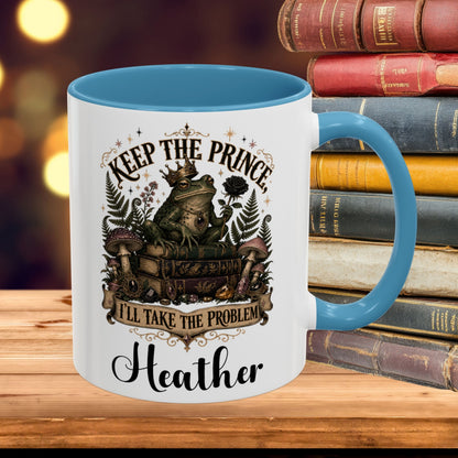 A white ceramic mug with a blue interior and handle, featuring a whimsical graphic of a frog wearing a crown and surrounded by mushrooms and books, with the text 'KEEP THE PRINCE I'LL TAKE THE PROBLEM' and 'Heather' below. The mug is placed on a wooden surface next to a stack of vintage books with a warm, blurred bokeh background.