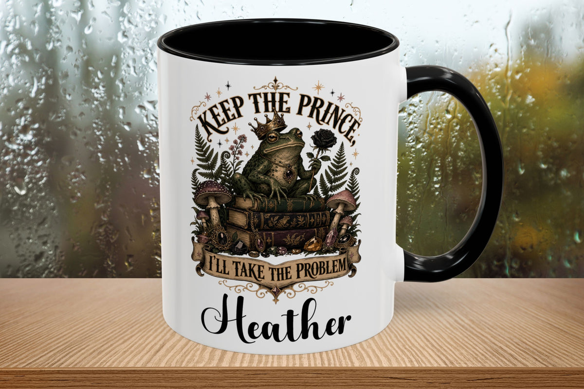 A white ceramic mug with a black interior and handle, featuring a whimsical graphic of a frog wearing a crown and the text 'KEEP THE PRINCE, I'LL TAKE THE PROBLEM' and 'Heather'. The mug sits on a wooden surface with a blurred, rainy window background.
