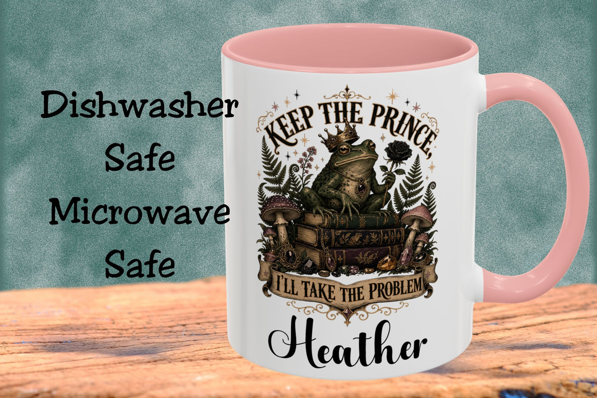 A white ceramic mug with a pink handle and interior, featuring a graphic of a frog wearing a crown over an open book with the text 'KEEP THE PRINCE I'LL TAKE THE PROBLEM' and the name 'Heather'. The mug is shown on a wooden surface against a textured teal background, with text on the left reading 'Dishwasher Safe Microwave Safe'.