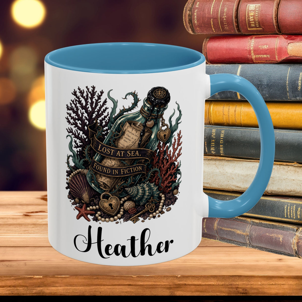 A white ceramic mug with a blue interior and handle, featuring a nautical illustration of a bottle at sea with the text 'LOST AT SEA FOUND IN FICTION' and the name 'Heather' printed below. The mug is styled on a wooden surface next to a stack of vintage books with a blurred warm background.