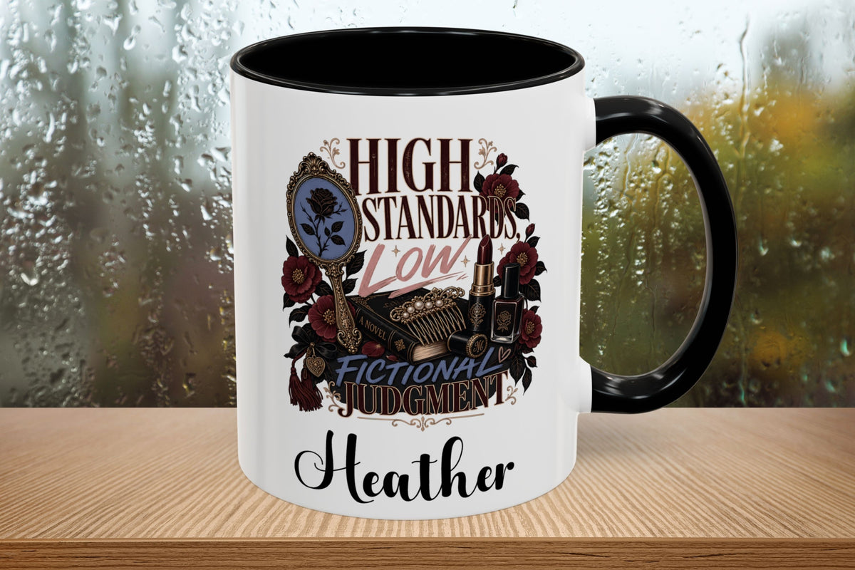 A white ceramic mug with a black handle and black interior, featuring a graphic design with roses, a mirror, and the text 'HIGH STANDARDS LOW FICTIONAL JUDGEMENT' and the name 'Heather' at the bottom. The mug sits on a wooden surface with a blurred, rainy window background.