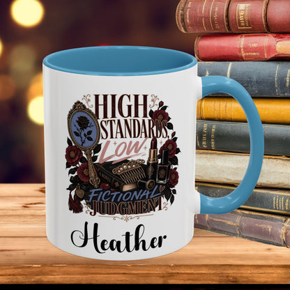A white ceramic mug with a blue interior and handle, featuring a decorative graphic that reads 'HIGH STANDARDS Low FICTIONAL JUDGMENT' with a mirror, roses, and lipstick illustration, plus the name 'Heather' printed below. The mug is placed on a wooden surface in front of a stack of vintage books with a blurred warm background.