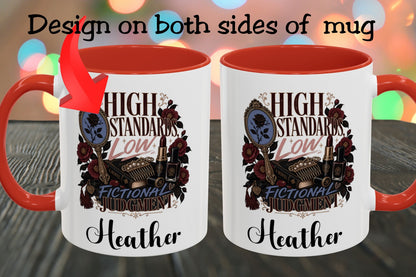 Two white ceramic mugs with orange-red interiors and handles are shown side by side on a dark wooden surface. The mugs feature a vintage-style graphic with the text 'HIGH STANDARDS LOW FICTIONAL JUDGEMENT' and the name 'Heather' printed below, plus a red arrow pointing to the left mug.