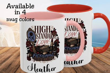Two white ceramic mugs with pink and orange-red interiors and handles, shown on a wooden surface with a blurred beach background. The mugs feature a decorative graphic with the words 'HIGH STANDARDS LOW FICTIONAL JUDGMENT' and the name 'Heather'.