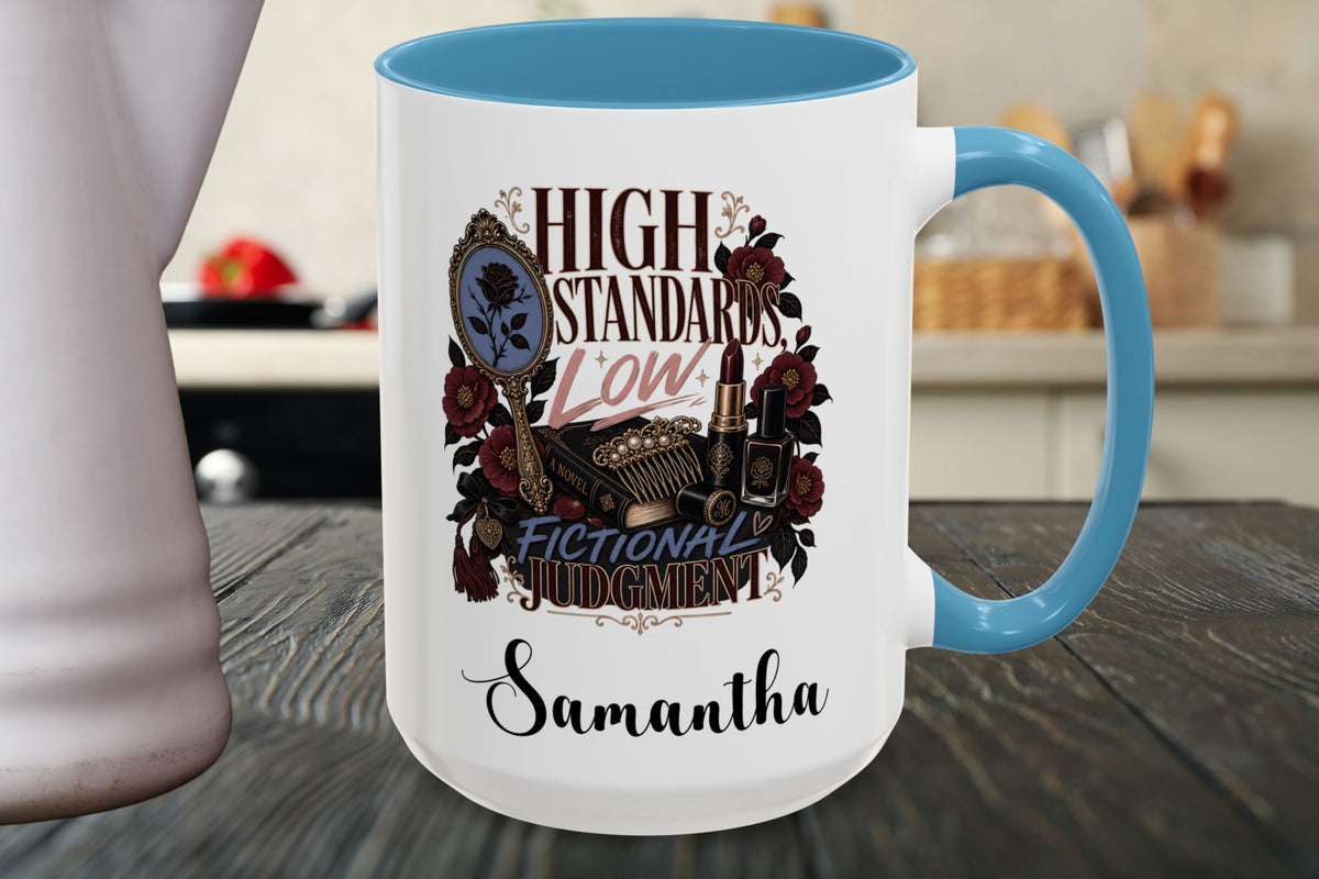 A white ceramic mug with a light blue handle and interior, featuring a decorative graphic with roses, a vintage camera, and the text 'HIGH STANDARDS LOW FICTIONAL JUDGMENT' above the name 'Samantha'. The mug is shown on a dark wooden table with a blurred kitchen background.