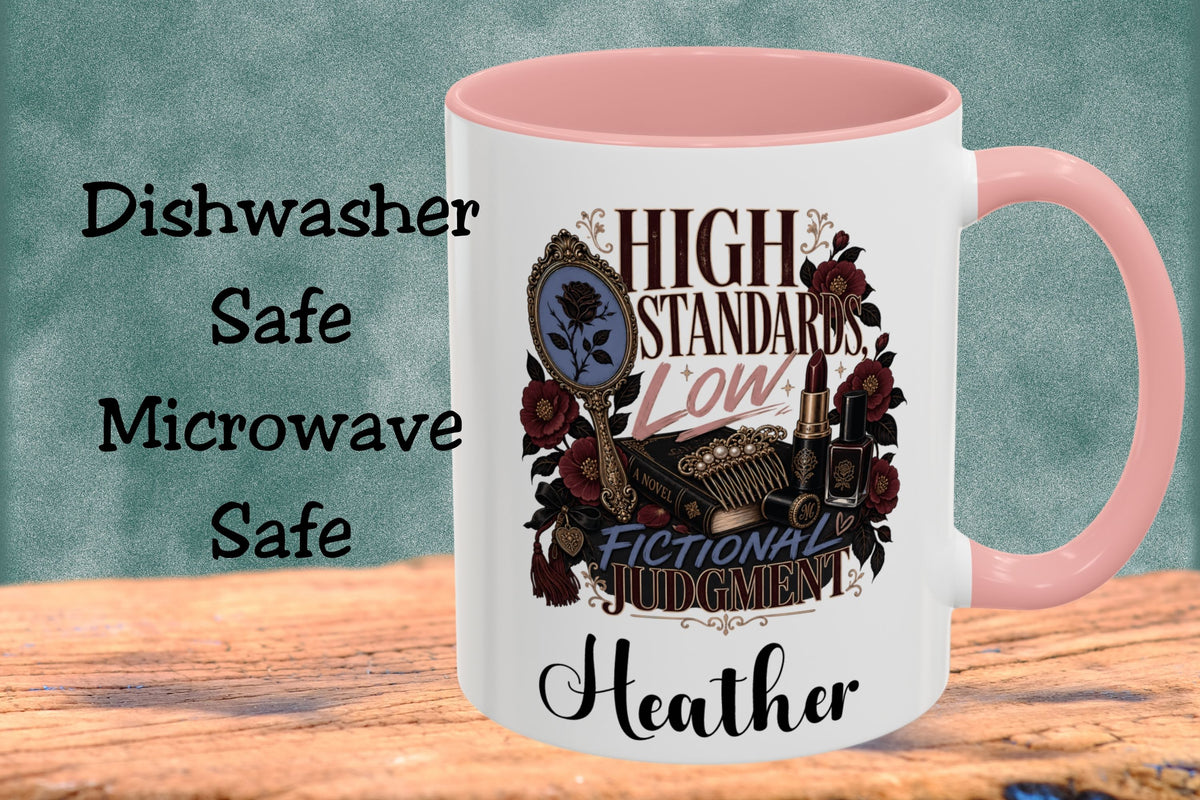 A white ceramic mug with a pink interior and handle, featuring a vintage-style graphic with the text 'HIGH STANDARDS LOW! FICTIONAL JUDGMENT' and the name 'Heather' at the bottom. The mug is shown on a wooden surface against a textured teal background with text on the left reading 'Dishwasher Safe Microwave Safe'.