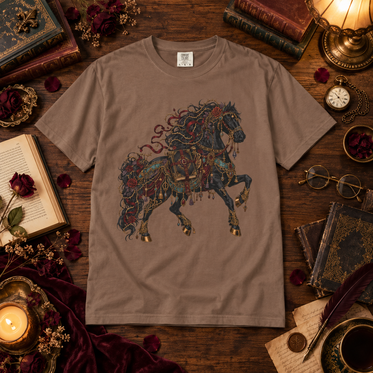 A taupe brown short-sleeve t-shirt featuring a detailed graphic of a rearing horse adorned with red roses and gold armor, displayed in a cozy, vintage-themed flat lay surrounded by old books, a candle, glasses, and a pocket watch.