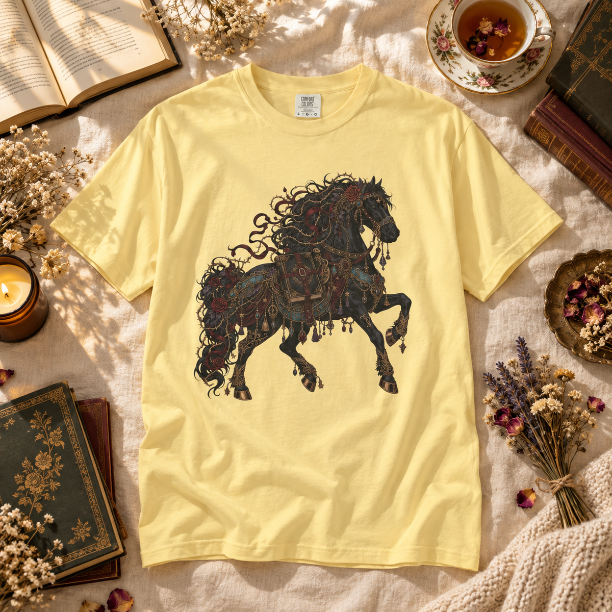 A pale yellow short-sleeve T-shirt laid flat on a textured cream fabric, featuring a large dark ornate horse graphic on the front. The shirt is styled in a cozy flat-lay arrangement with books, dried flowers, a cup of tea, and a candle.
