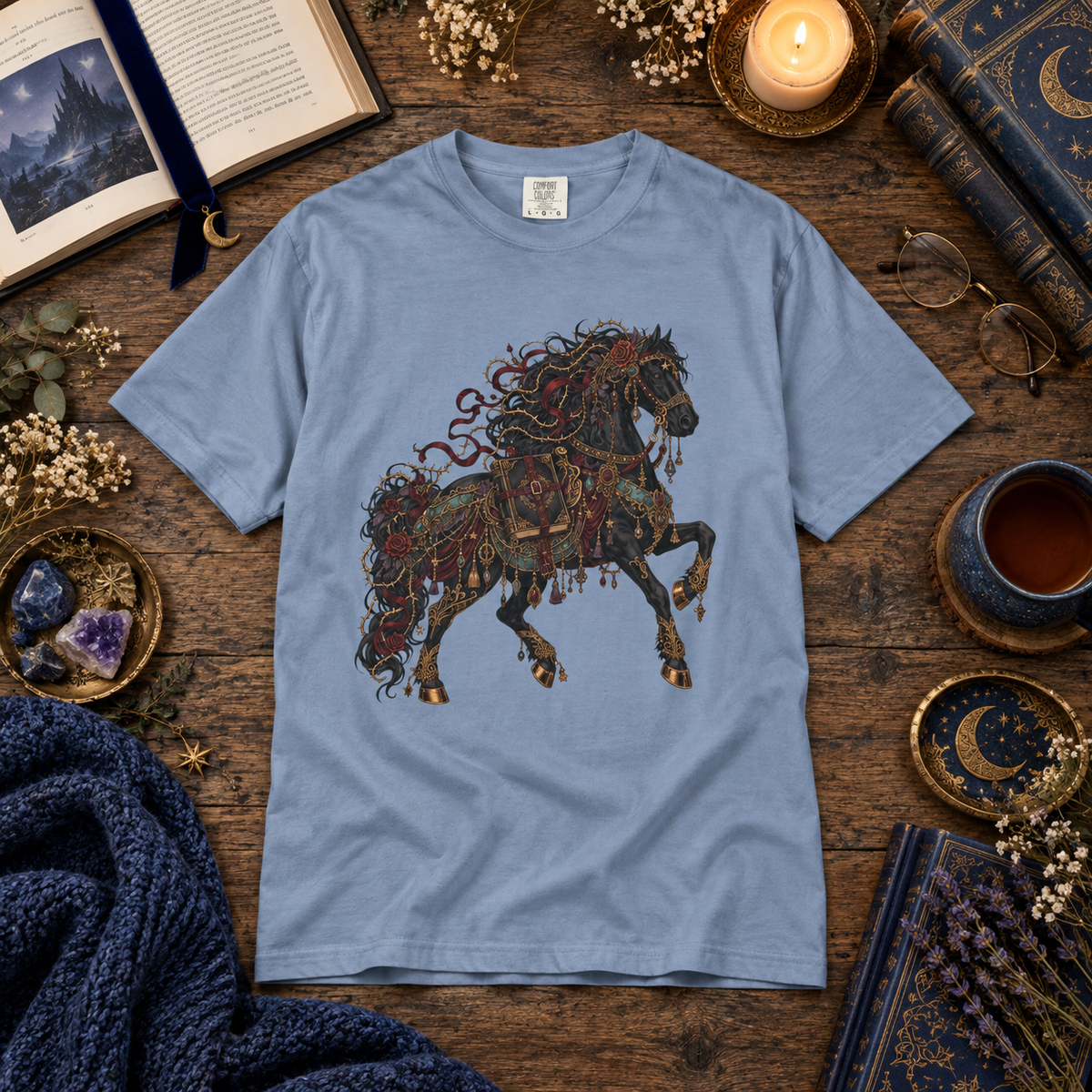 A light blue crewneck t-shirt featuring a detailed graphic of a dark horse adorned with red and gold snakes, laid flat on a rustic wooden table surrounded by cozy books, candles, and dried flowers.