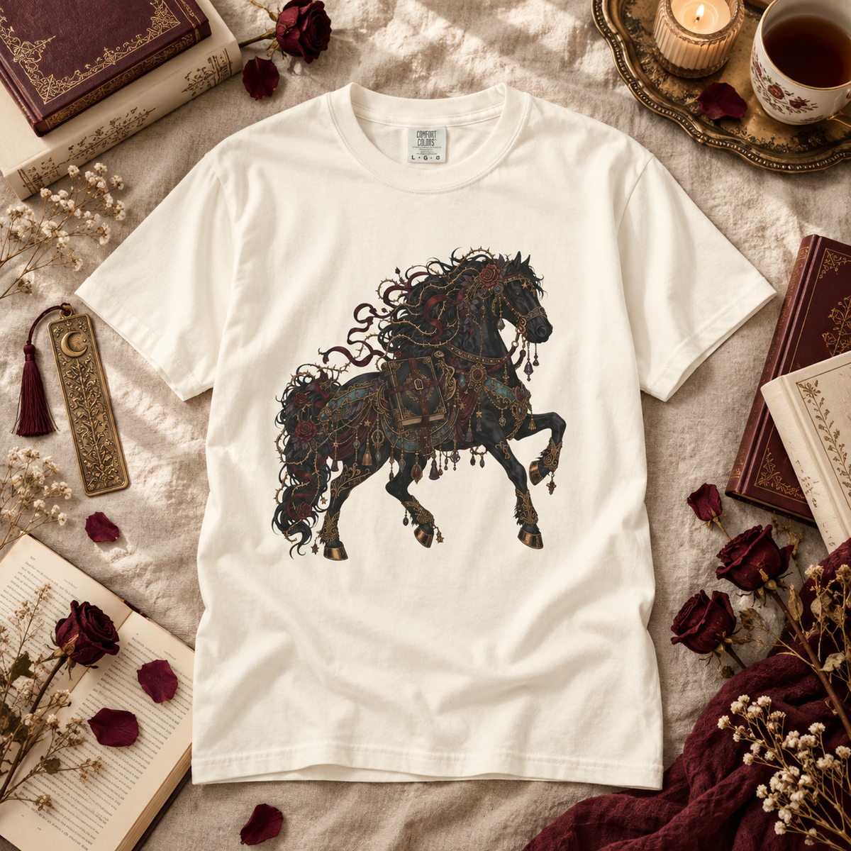 An off-white short-sleeve t-shirt featuring a detailed graphic of a dark, ornate horse with gothic and filigree details, styled in a cozy flat-lay arrangement with books, dried roses, and a candle.