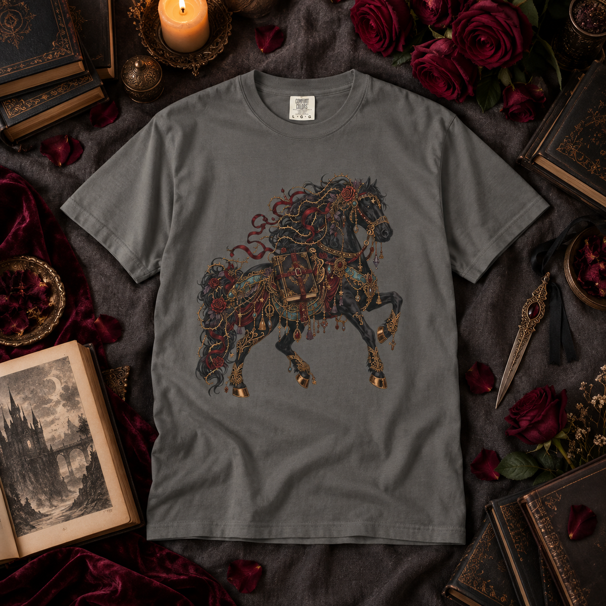 A dark gray crewneck t-shirt featuring a detailed graphic of a rearing horse adorned with ornate gold, red, and black filigree patterns. The shirt is presented in a styled flat-lay arrangement surrounded by gothic-themed props including vintage books, red roses, velvet fabric, and a lit candle.