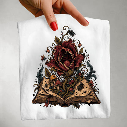 A hand with red-painted nails holds a white t-shirt featuring a detailed graphic of a monstrous red flower rising from an open book, surrounded by mushrooms, insects, and dark swirling details, set against a neutral gray background.