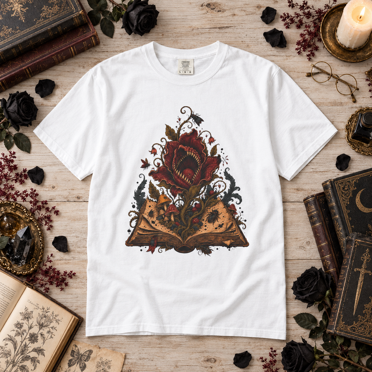 A white short-sleeve graphic T-shirt laid flat on a rustic wooden surface, surrounded by vintage books, black roses, dried flowers, glasses, and a candle. The shirt features a large central graphic of an open book with a red rose and dark ornate details.