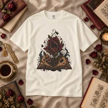 A white short-sleeve crewneck t-shirt with a detailed graphic of an open book and a red rose, styled in a flat lay on a beige linen fabric surrounded by vintage books, dried roses, a teacup, and a candle.