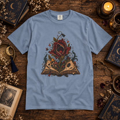 A light blue crewneck t-shirt with a detailed graphic of a giant red flower blooming from an open book, surrounded by vintage books, glasses, and a candle on a dark wooden table.