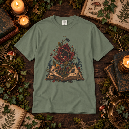A sage green short-sleeve t-shirt with a detailed graphic of a red rose growing from an open book, surrounded by fantasy elements like a dragon, on a dark wood background styled with books, candles, moss, and ivy.