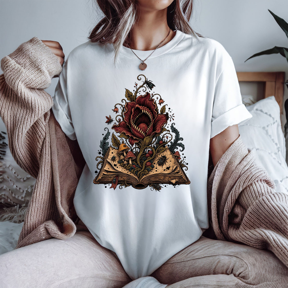 A woman wearing a white t-shirt with a graphic of a giant red flower growing from an open book, styled with a beige knit sweater draped off her shoulders in a cozy bedroom setting.