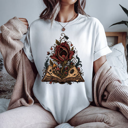 A woman wearing a white t-shirt with a graphic of a giant red flower growing from an open book, styled with a beige knit sweater draped off her shoulders in a cozy bedroom setting.