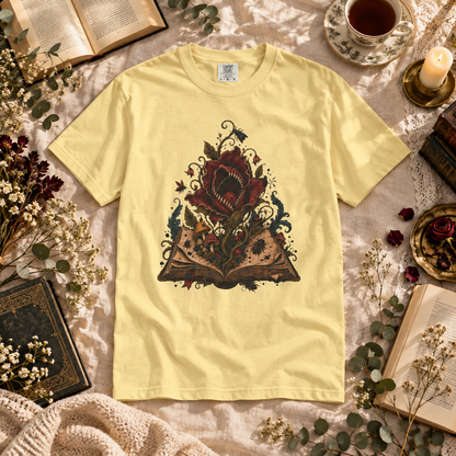 A pale yellow graphic t-shirt laid flat on a textured beige fabric, surrounded by vintage-style props including books, dried flowers, a teacup, and a candle. The t-shirt features a detailed graphic of a red rose with a bird inside its petals, growing from an open book.