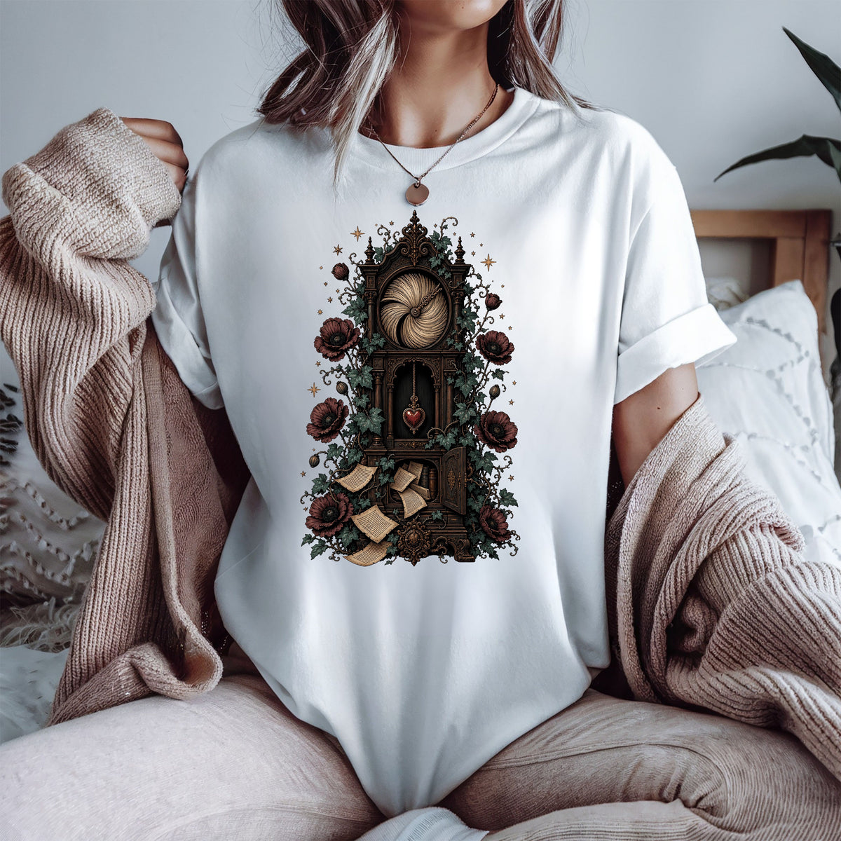 A woman wearing a white t-shirt with a detailed gothic illustration of a clockwork mechanism, surrounded by roses and scrolls, styled with a beige knit cardigan in a cozy bedroom setting.