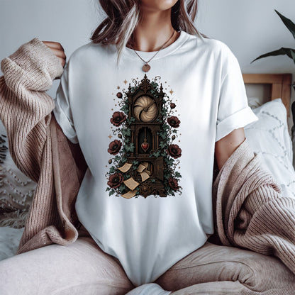 A woman wearing a white t-shirt with a detailed gothic illustration of a clockwork mechanism, surrounded by roses and scrolls, styled with a beige knit cardigan in a cozy bedroom setting.