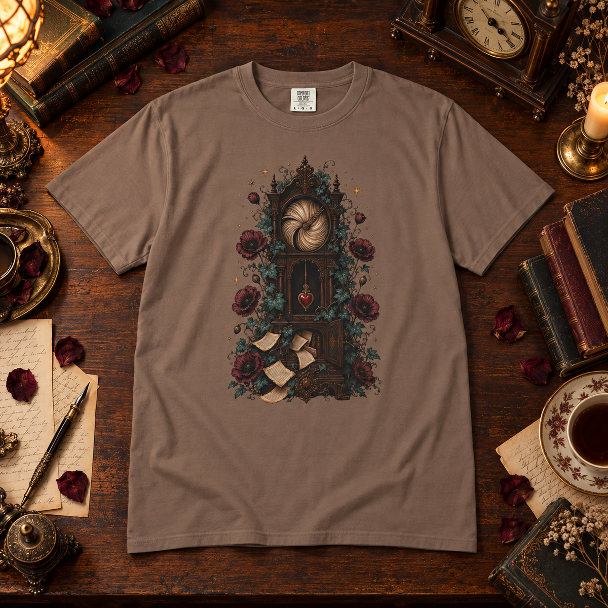 A taupe brown short-sleeve t-shirt with a detailed gothic clock graphic, laid flat on a dark wooden table surrounded by vintage props like books, a clock, and a cup of tea.