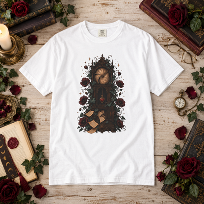 A white short-sleeve crewneck t-shirt with a dark fantasy illustration of a bookshelf, roses, and books, presented in a flat-lay setting on a rustic wooden table surrounded by decorative props.