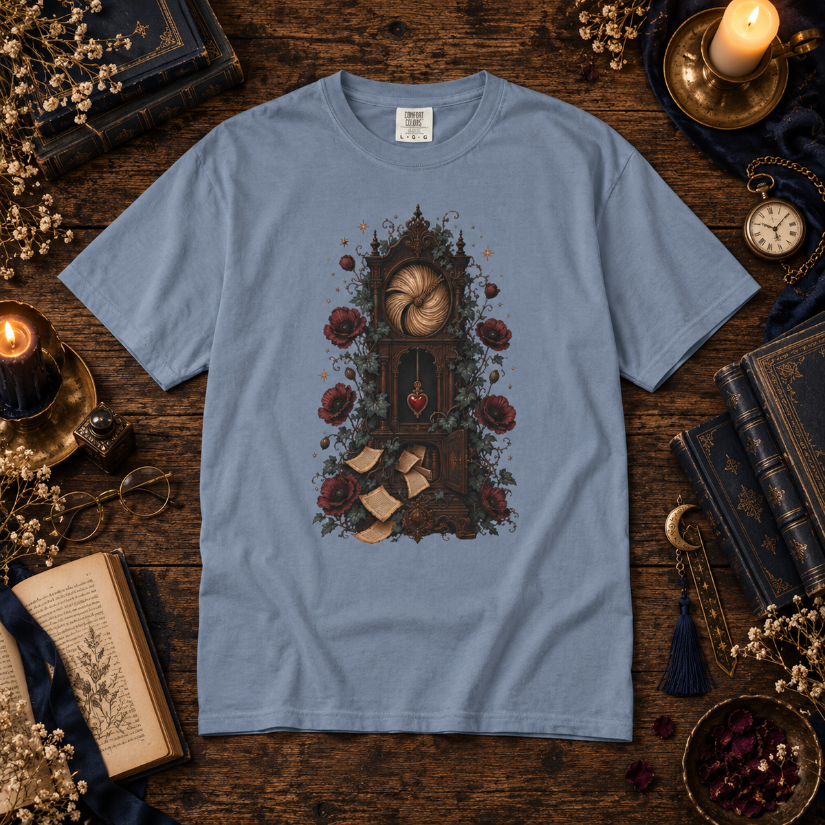 A light blue crewneck t-shirt with a detailed gothic illustration of a grandfather clock surrounded by roses and scrolls, presented in a flat-lay on a dark wooden table surrounded by vintage props like candles, books, and a pocket watch.