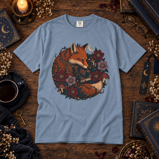 A light blue crewneck t-shirt featuring a detailed graphic of a fox surrounded by flowers and a key, laid flat on a rustic wooden table surrounded by cozy props like a mug, glasses, and old books.