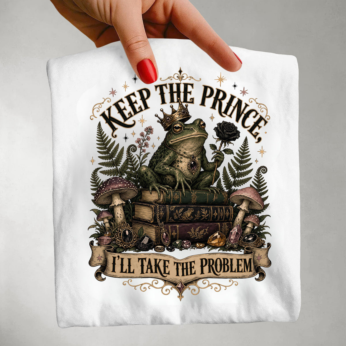 A hand with red nail polish holds up a white t-shirt featuring a whimsical graphic of a crowned frog sitting on books, surrounded by mushrooms, ferns, and a rose, with the text 'KEEP THE PRINCE, I'LL TAKE THE PROBLEM'.